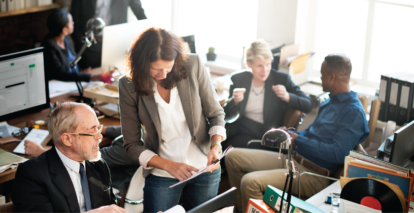 A woman stands at a desk, engaged in discussion with several colleagues around her.