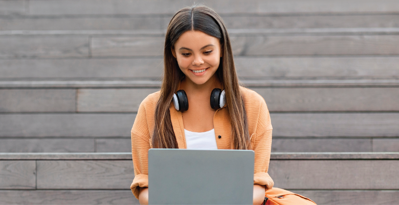 A woman wearing headphones sits on steps, focused on her laptop, with a casual outdoor setting in the background.