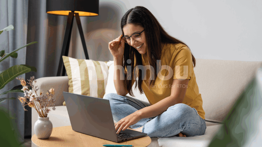 A young woman sits on a couch, focused on her laptop, with a cozy living room setting in the background.